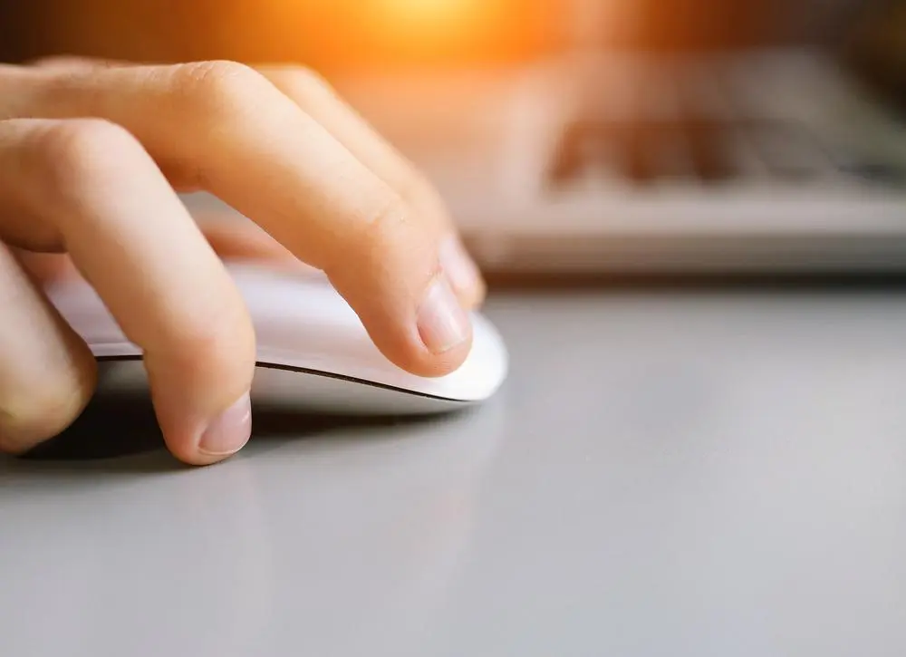 A close-up of a persons hand using a white computer mouse on a gray surface. In the background, a laptop keyboard is partially visible. Warm sunlight creates a bright, glowing effect from above.