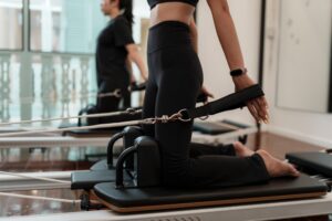 Two women kneel on reformer machines in a Pilates studio, holding resistance straps. Both wear black athletic wear and appear focused on their workout. The room is bright with large windows in the background.
