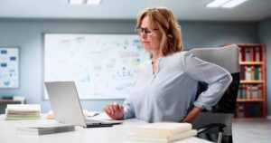 A woman sitting at a desk with a laptop, holding her lower back and wincing in pain, suggesting discomfort or back pain. Books and office supplies are on the desk, and bookshelves are in the background.