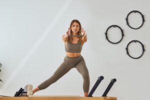 A woman in athletic wear performs a stretching exercise with a resistance band, standing on a Pilates reformer. Three Pilates rings are mounted on the white wall behind her.
