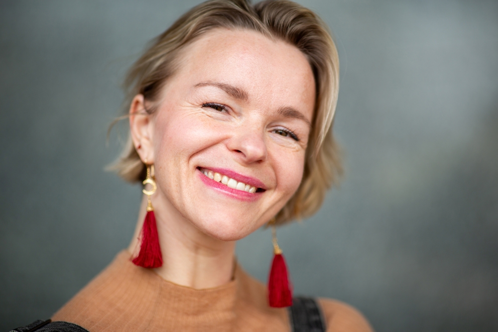 A smiling woman with short blonde hair wears red tassel earrings and a beige top, posing in front of a blurred gray background.