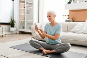 Smiling older woman with short gray hair sits cross-legged on a yoga mat in a living room, looking at and using a smartphone, with a sofa and plants in the background.